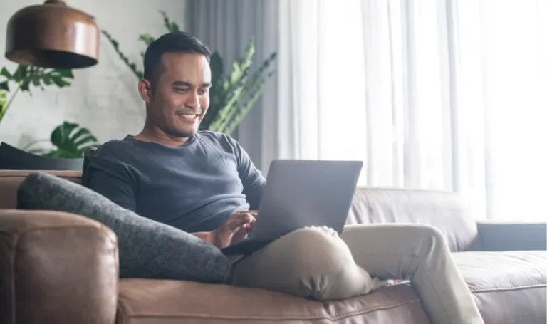 A man is sitting on his couch as he uses his laptop.